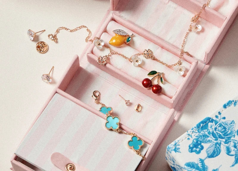 An open pink striped jewelry box displayed on a light beige surface, containing multiple delicate gold-tone jewelry pieces including earrings, necklaces, and bracelets with turquoise, red, yellow, and white accents. A blue-and-white patterned box is partially visible at the lower right.